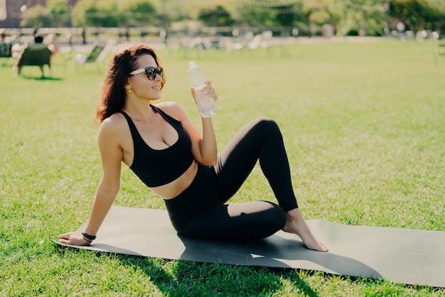 Foto ao ar livre de mulher magra ativa bebe água da garrafa sente sede após o treinamento físico concentrado na distância usa top recortado e legging pratica ioga fora do conceito de esporte