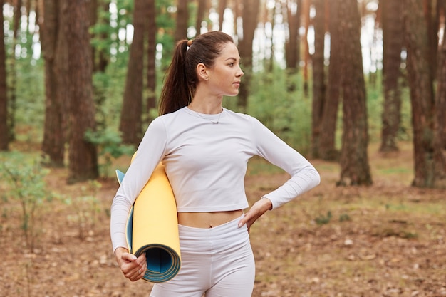 Foto ao ar livre de mulher magra e confiante em roupas esportivas brancas, segurando o tapete de ioga nas mãos, olhando para longe e mantendo as mãos nos quadris