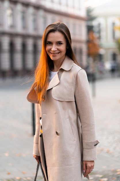 Foto ao ar livre de senhora morena andando na rua, fundo em dia de outono. retrato de estilo de rua da moda. vestindo calças escuras casuais, suéter branco e casaco cremoso. conceito de moda.