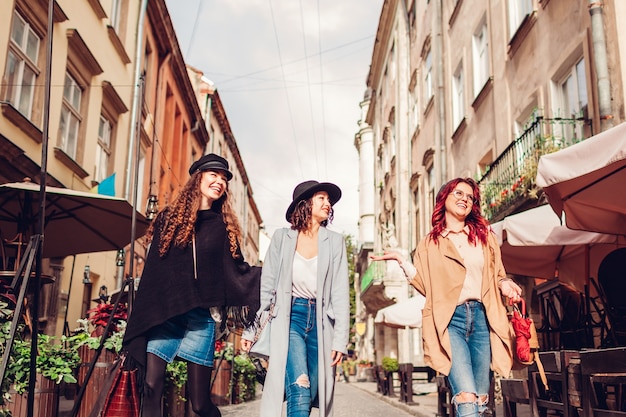 Foto ao ar livre de três mulheres caminhando na rua da cidade. Garotas felizes conversando e se divertindo