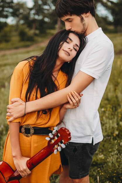 Foto ao ar livre de um casal romântico abraçando ao ar livre. linda jovem morena segurando uma guitarra inclinada para a cabeça com os olhos fechados no peito do namorado.