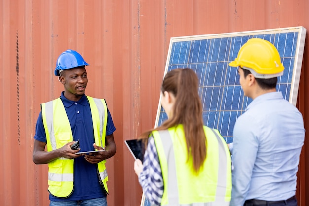 Foto ao ar livre de um engenheiro negro africano inspecionando painel solar elétrico usando capacete, óculos de proteção e equipamento de segurança,