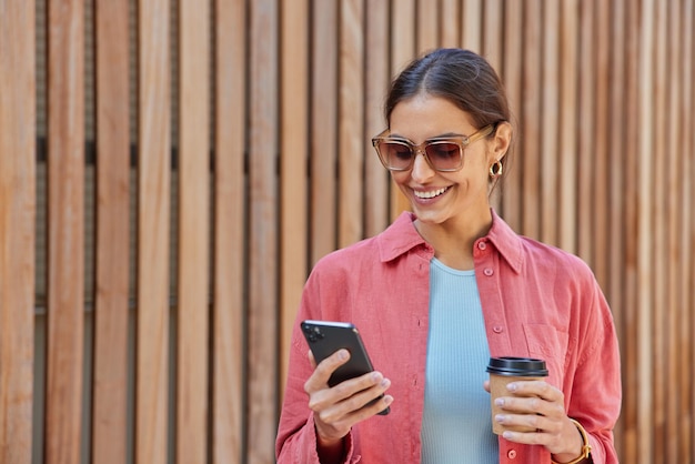 Foto ao ar livre de uma mulher europeia bonita morena usa telefone celular para comunicação on-line e bebe café para viagem usa óculos de sol da moda camisa rosa posa contra madeira, portanto, se sente feliz