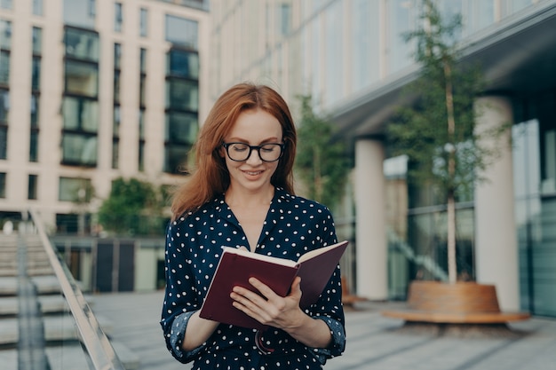 Foto ao ar livre de uma mulher ruiva fazendo anotações no bloco de notas e planejando o dia de trabalho