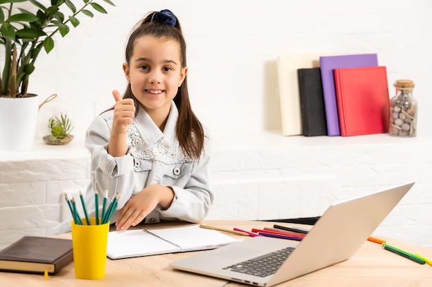 Foto aproximada da aluna bonita da escola fala skype notebook lição on-line videochamada sentar mesa usar fones de ouvido distância cumprimentar colegas onda braço quarentena estudar sala de estar dentro de casa