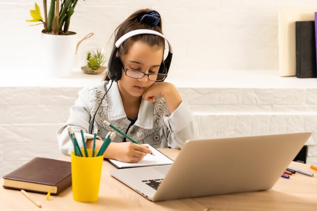 Foto foto aproximada da aluna bonita da escola fala skype notebook lição on-line videochamada sentar mesa usar fones de ouvido distância cumprimentar colegas onda braço quarentena estudar sala de estar dentro de casa