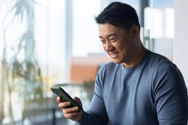 Foto aproximada de empresário masculino usando telefone no escritório moderno asiático olhando para a tela do telefone e sorrindo chamada on-line