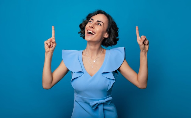 Foto aproximada de uma jovem linda e feliz em um vestido azul que está apontando com o dedo indicador e isolada em fundo azul