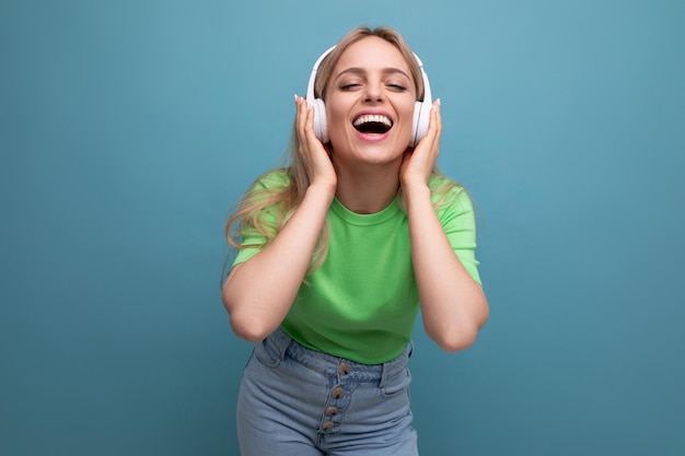 Foto aproximada de uma loira estilosa com uma roupa casual cantarolando uma música em grandes fones de ouvido brancos em um