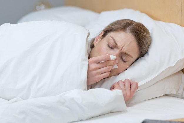 Foto aproximada de uma mulher doente deitada debaixo de um cobertor na cama em casa tem um nariz escorrendo e frio