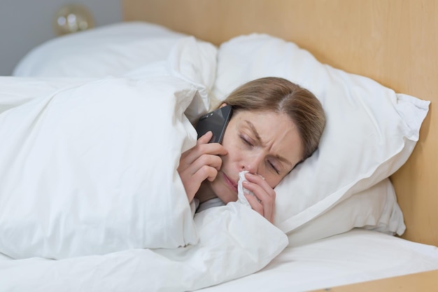 Foto aproximada de uma mulher doente em casa tem um nariz escorrendo ruim e está frio na cama coberta com um cobertor chama um médico para aconselhamento