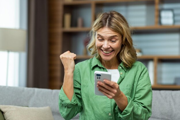 Foto aproximada, uma mulher idosa feliz está sentada no sofá em casa, segurando o telefone nas mãos