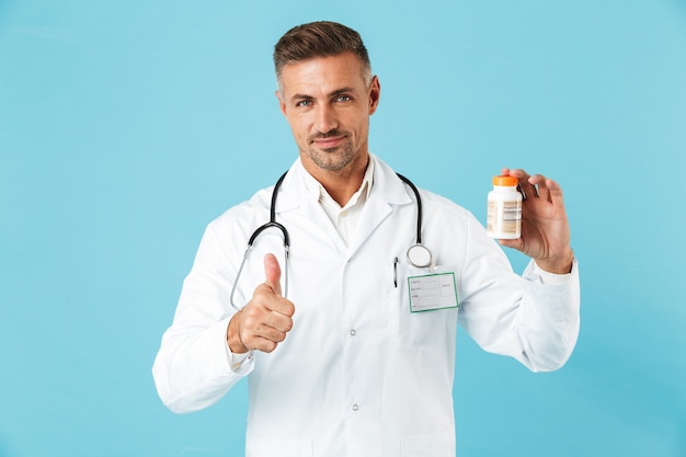 Foto de un apuesto hombre maduro médico posando aislado sobre la pared de la pared azul con píldoras vitaminas mostrando los pulgares para arriba.