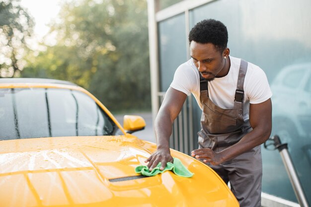 Una foto de un apuesto joven africano barbudo que limpia el capó de un auto amarillo