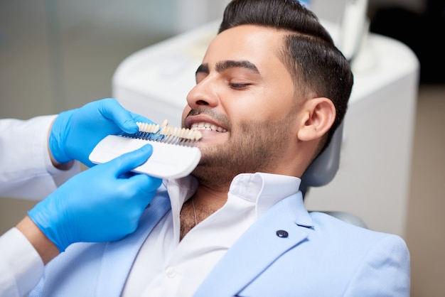 Foto de un apuesto joven en una cita médica en la oficina del dentista dentista profesional sosteniendo dentaduras postizas comparando el color y el tono del blanqueamiento de la salud de la medicina dental.