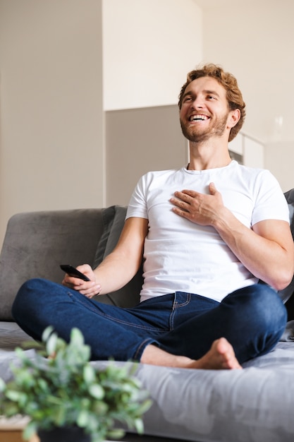 Foto de un apuesto joven feliz en el sofá en casa con control remoto ver televisión.