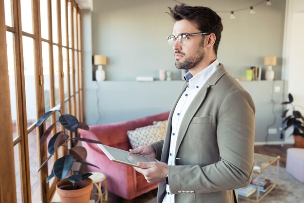 Foto de apuesto joven hombre de negocios mirando hacia los lados mientras usa su tableta digital en el pasillo de la oficina.