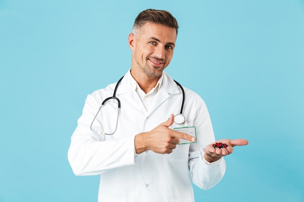 Foto de un apuesto médico hombre maduro posando aislado sobre pared azul con píldoras vitaminas.