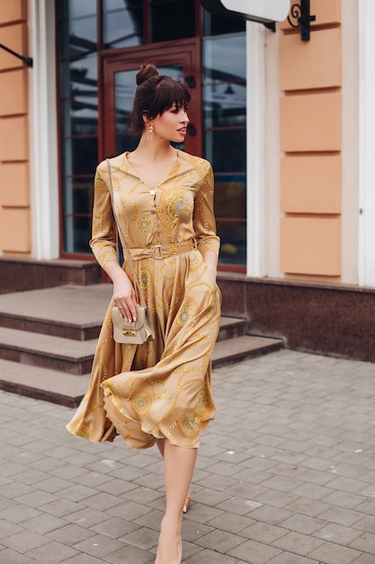 Foto de archivo de cuerpo entero de una hermosa dama elegante en vestido dorado, tacones altos de color dorado y bolso cruzado. Ella camina en la calle sonriendo.