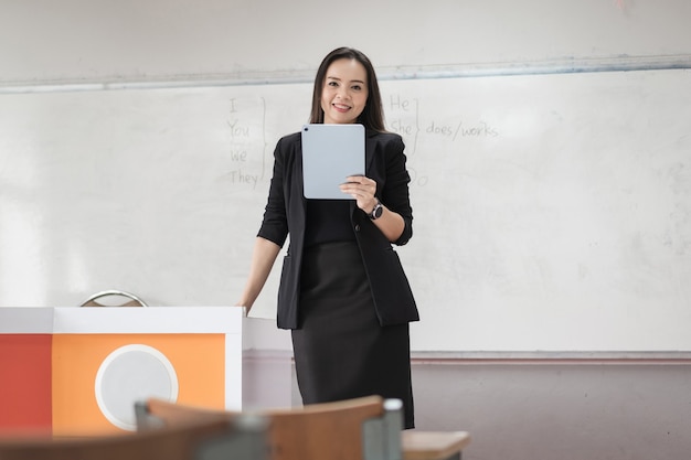 Foto de archivo retrato de una profesora asiática alegre confiada en un uniforme de traje negro con una tableta digital y una computadora portátil para enseñar el idioma moderno en el aula