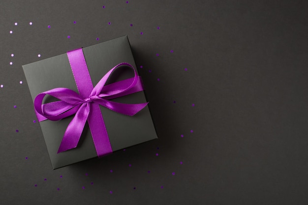 Foto de arriba de caja de regalo negra con cinta violeta envuelta como arco y confeti alrededor aislado en el fondo negro