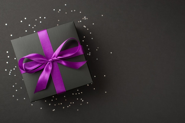 Foto de arriba de una caja de regalo negra con una cinta violeta envuelta como confeti de arco y brillantina alrededor aislada en el fondo negro