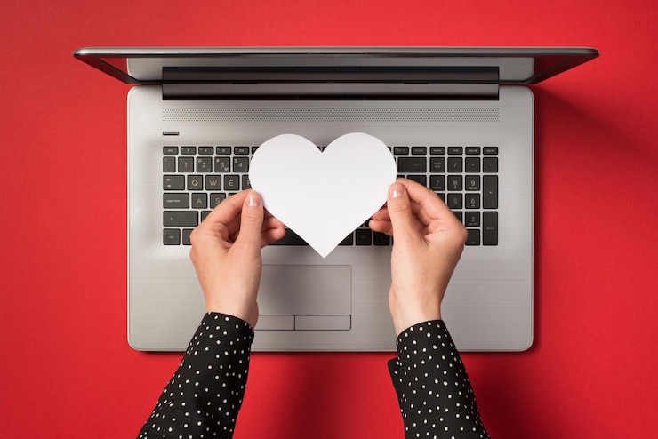 Foto foto de arriba de una computadora portátil gris y manos sosteniendo un corazón de papel blanco aislado en el fondo rojo