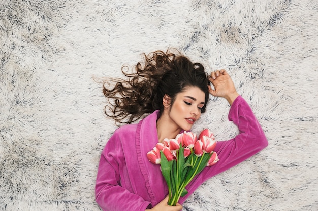 Foto de arriba de hermosa mujer con pelo largo y rizado vistiendo bata de niña sosteniendo flores mientras está acostada sobre la piel blanca en el apartamento