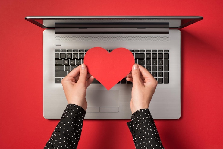 Foto foto de arriba de una laptop gris y manos sosteniendo un corazón de papel rojo aislado en el fondo rojo