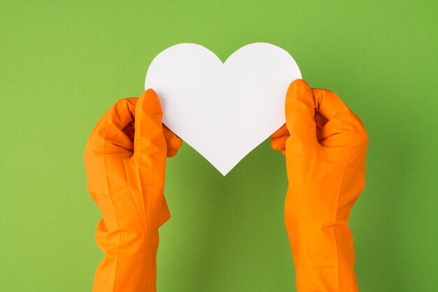 Foto de arriba de manos con guantes naranjas sosteniendo la tarjeta de papel blanco como corazón aislado en el fondo verde