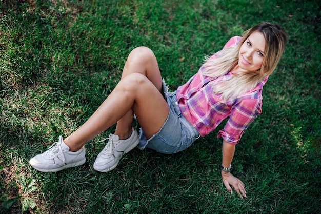 Foto desde arriba de una mujer rubia brillante adulta sentada en la hierba en el parque con las manos en la hierba usando una falda de jeans de camisa a cuadros y zapatillas blancas durante el día Fondo adorable lleno de verde