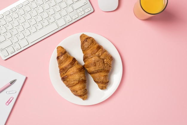 Foto de arriba del teclado, computadora, mouse, vaso de jugo de naranja, bloc de notas con clips de papel y plato con dos croissants aislados en el fondo rosa