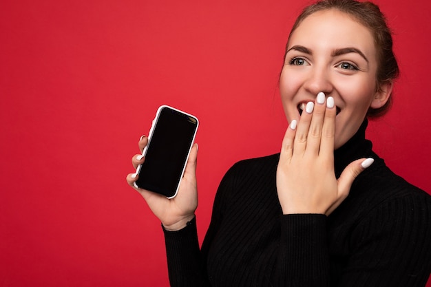 Foto de asombrada atractiva joven morena positiva vistiendo un suéter negro que se encuentran aisladas sobre la pared roja que muestra el teléfono móvil con pantalla vacía