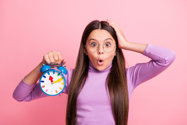 Foto de asombrado niño pequeño niña mantenga el reloj toque la cabeza de la mano aislada sobre fondo de color pastel