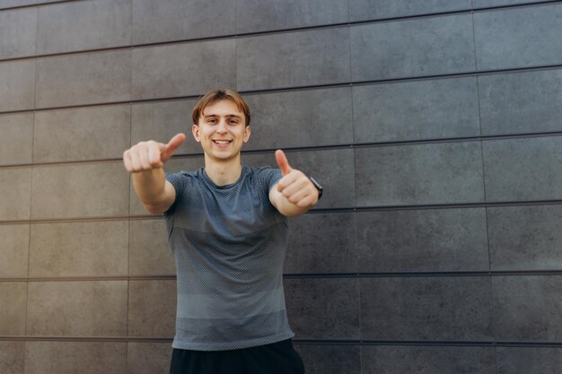 Foto de un atleta fuerte que se encuentra aislado sobre un fondo de pared gris Mirando a la cámara apuntando con el pulgar hacia arriba, un tipo alegre y emocionado