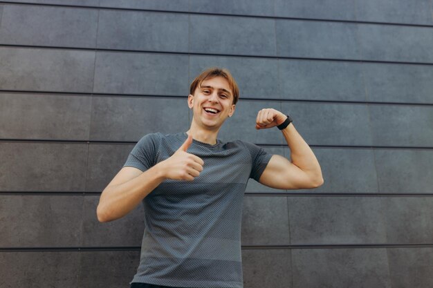 Foto de un atleta fuerte que se encuentra aislado sobre un fondo de pared gris Mirando a la cámara apuntando con el pulgar hacia arriba, un tipo alegre y emocionado