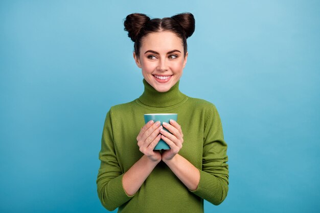 Foto de atractiva dama adolescente encantadora sostener bebida caliente taza té café disfrutar fin de semana mañana mirar lado espacio vacío usar cálido verde jersey de cuello alto aislado pared de color azul