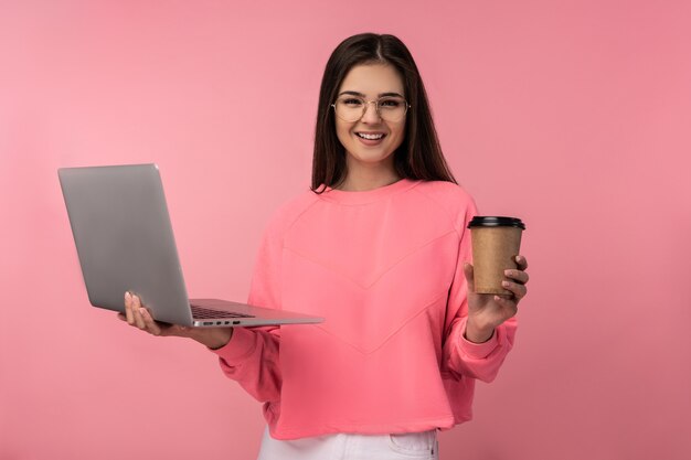 Foto de una atractiva dama con gafas sonríe, sostiene un café portátil y trabaja en línea