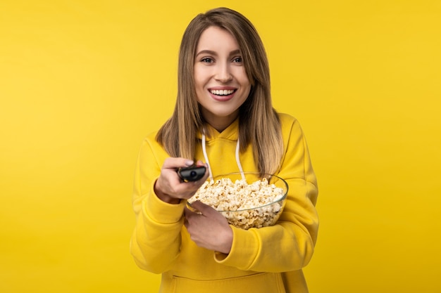 La foto de la atractiva dama sostiene el control remoto de la televisión y un plato de palomitas de maíz, con cara feliz intenta encender un canal. Viste con capucha amarilla casual, fondo de color amarillo aislado.