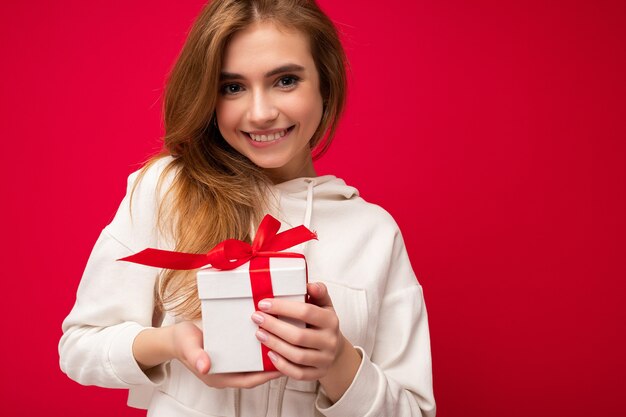 Foto de atractiva feliz sonriente linda rubia joven mujer aislada sobre fondo rojo pared vistiendo sudadera con capucha blanca con caja de regalo blanca con cinta roja y mirando a cámara. Espacio libre