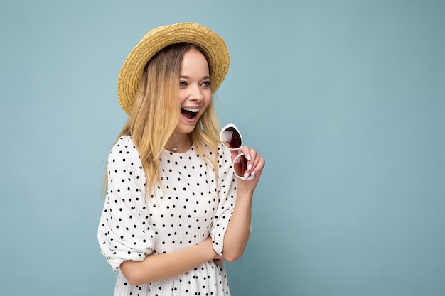 Foto de una atractiva joven rubia sonriente feliz vistiendo ropa casual de verano y elegantes gafas de sol aisladas sobre la pared de colores de fondo mirando hacia el lado