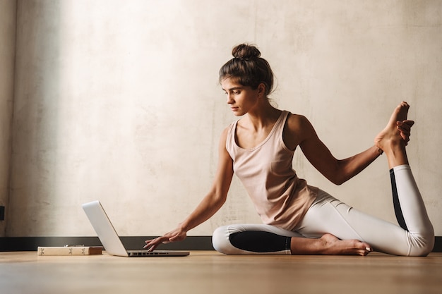 Foto foto de atractiva mujer concentrada en ropa deportiva haciendo ejercicios de yoga usando la computadora portátil mientras está sentado en el piso en casa