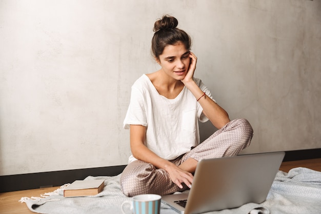 Foto de atractiva mujer concentrada vistiendo ropa de ocio tomando café y usando la computadora portátil mientras está sentado en el piso en casa