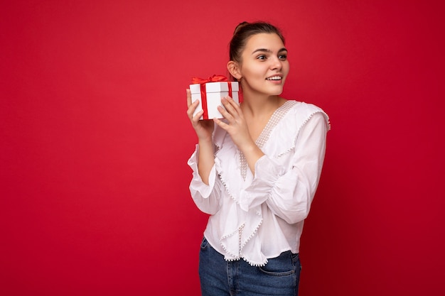 Foto de atractiva mujer morena joven sonriente positiva aislada sobre fondo rojo pared vistiendo blusa blanca sosteniendo caja de regalo blanca con cinta roja y mirando hacia el lado.