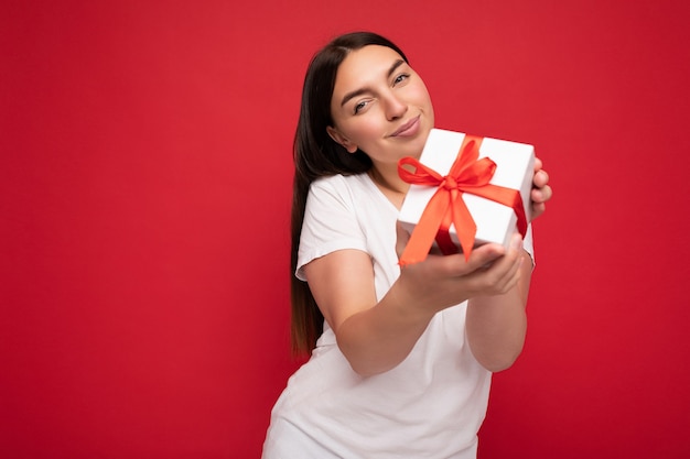 Foto de atractiva mujer morena joven sonriente positiva aislada sobre la pared de colores de fondo vistiendo ropa de moda todos los días con caja de regalo y mirando a la cámara.