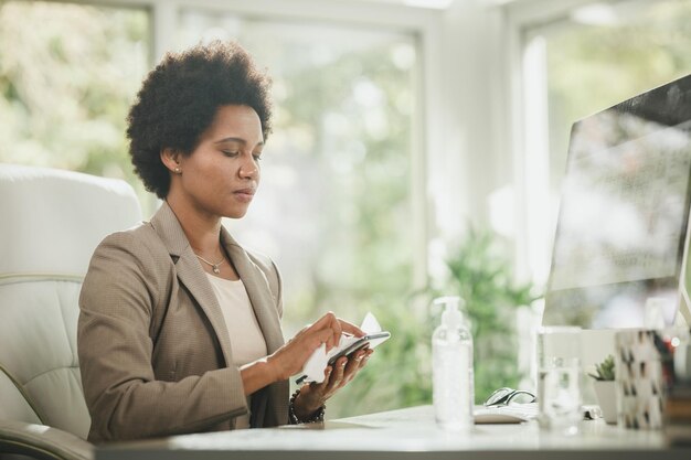 Foto de una atractiva mujer de negocios africana sentada sola en su oficina y usando gel antiséptico para desinfectar su smartphone durante la pandemia de COVID-19.