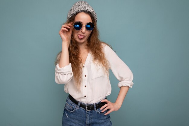 Foto de atractiva mujer rizada joven feliz aislado sobre fondo azul vistiendo una blusa blanca casual y elegantes gafas mirando a la cámara y divirtiéndose