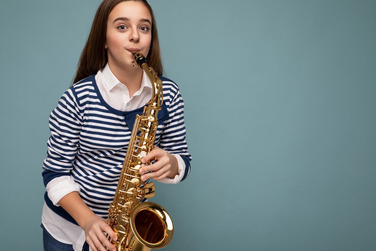 Foto foto de la atractiva niña brunet feliz positiva vistiendo elegante manga larga a rayas que se encuentran aisladas sobre fondo azul pared tocando el saxofón mirando a la cámara. espacio vacio