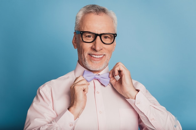 Foto de atractivo hombre de edad ajustando la pajarita aislado sobre fondo azul.