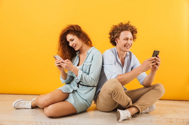 Foto de atractivo hombre y mujer caucásicos sentados en el suelo espalda con espalda y usando teléfonos inteligentes, aislado sobre fondo amarillo